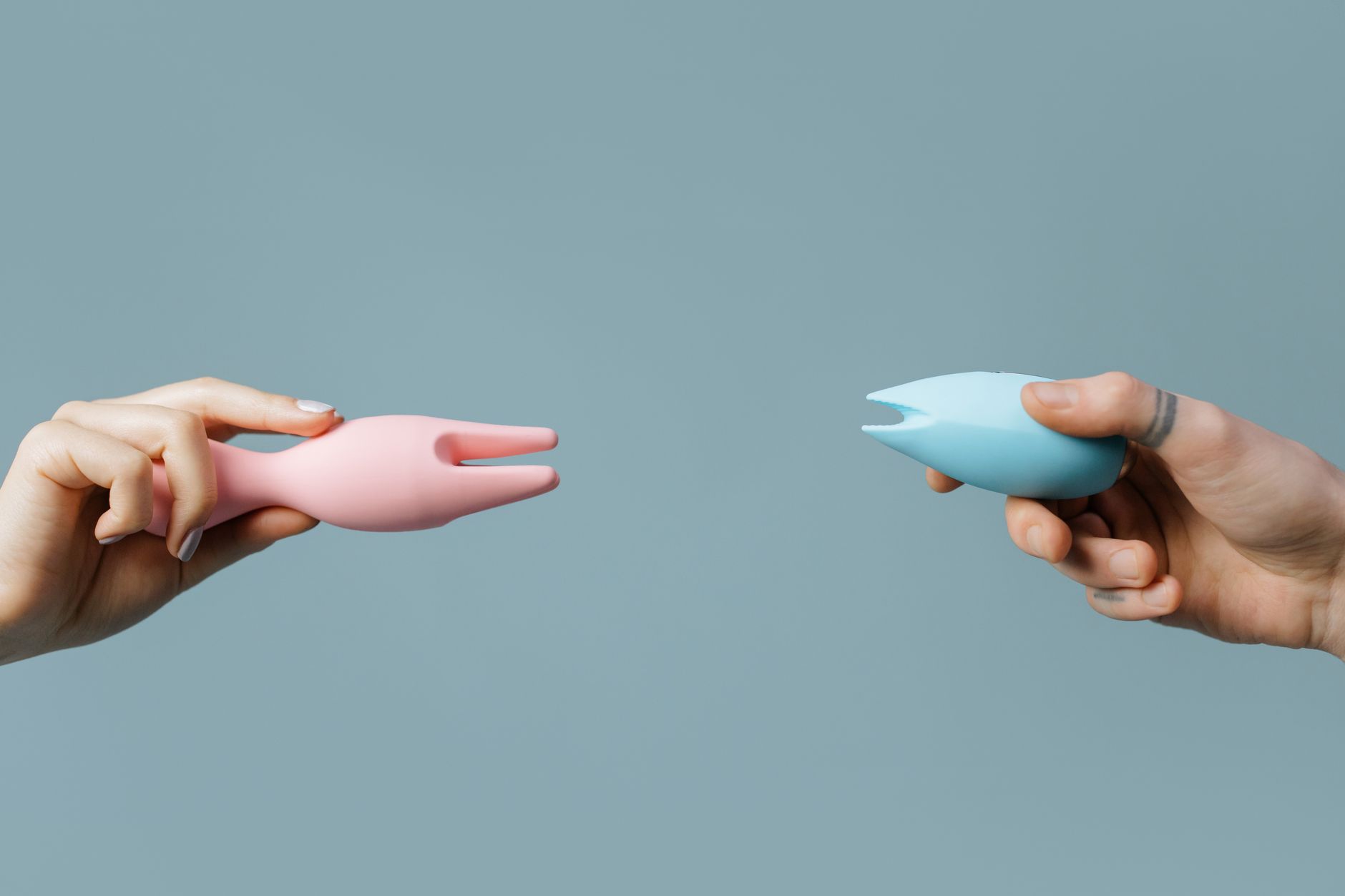 Two hands holding pink and blue lemon clitoral vibrators against a soft pastel background, symbolizing accessible pleasure options.