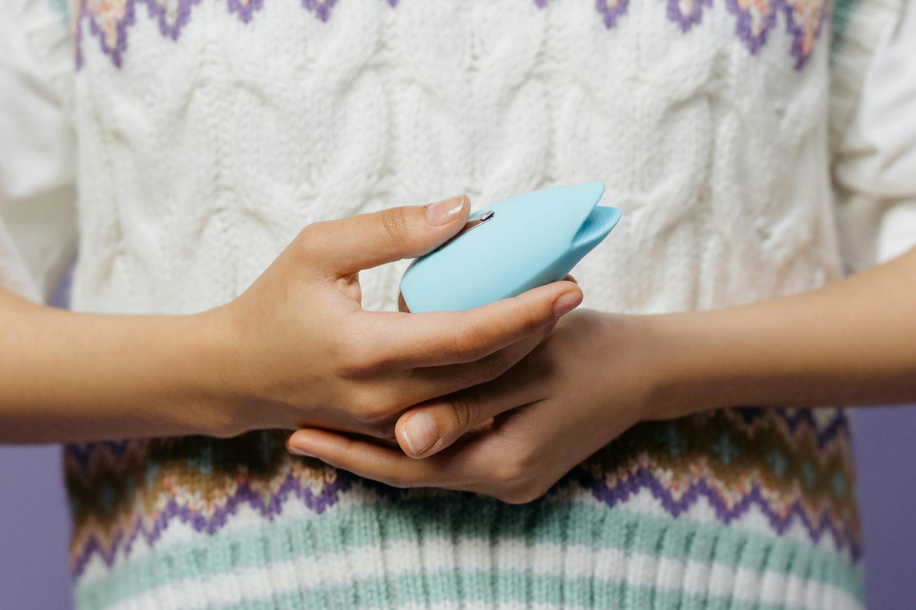 Detalhe de mãos segurando um vibrador azul contra uma blusa tricotada, pronto para reconectar com o prazer.